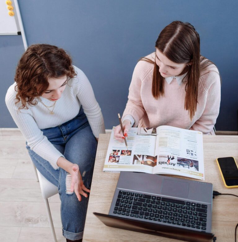 Two women discussing plans in an office using a laptop and magazine
